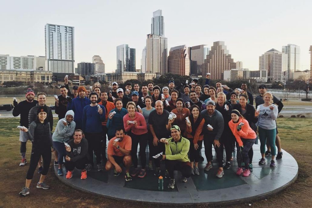 Image of group of runners posing in front of the Austin skyline holding Tacodeli tacos. Tacodeli is the Official Taco of the Austin Marathon.