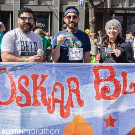 Image of two participants showing off their medals to a friend while they enjoy beers at the Oskar Blues beer garden at the 2019 Austin Marathon, capping off an unforgettable weekend.