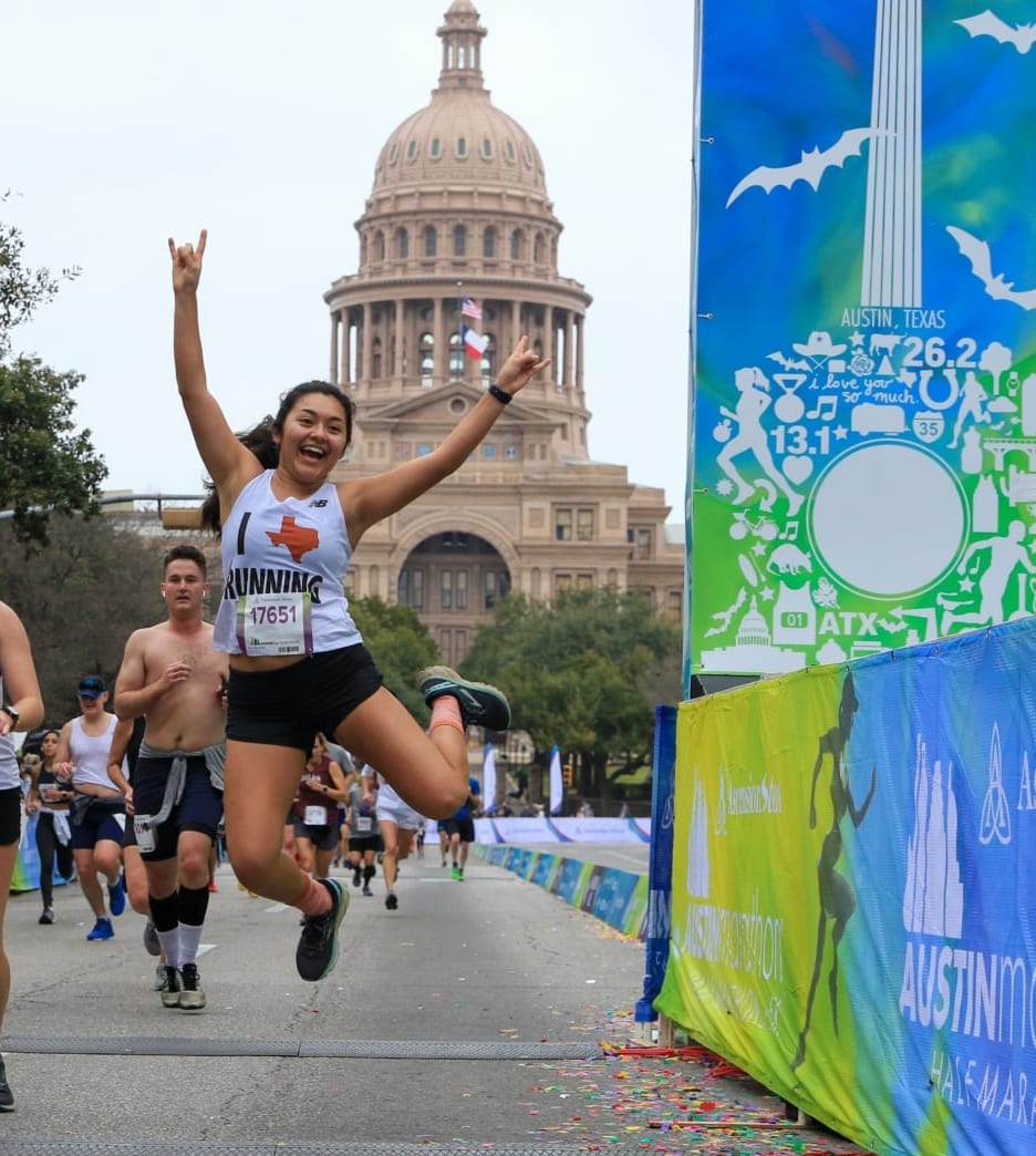 Image of runner jumping while crossing the 2020 Austin Half Marathon finish line. She's in mid-air making the Longhorn sign with both hands. Read this blog and learn how to gain strength so you can cross the finish line like this!