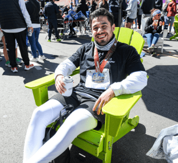 A man wearing a black long-sleeve shirt, white leggings, and a race medal is sitting on a bright green Adirondack chair. Smiling and holding a drink can, he relaxes among other runners in the background, all enjoying a post-race event celebrating their performance at the Austin Marathon Half Marathon & 5K