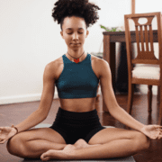 A person with curly hair sits cross-legged on a yoga mat, eyes closed and hands resting on their knees in a meditation pose. Wearing a blue crop top and black shorts, they exemplify pushing through life's challenges. The background includes a wooden table and chair. Austin Marathon Half Marathon & 5K