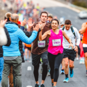 Runners in the marathon push through and exchange high-fives with spectators along the course. A woman in a pink top smiles while engaging with the crowd, embodying the spirit of conquering hard miles. Other runners follow behind, their colorful athletic wear visible against the blurred background that enhances the sense of movement. Austin Marathon Half Marathon & 5K