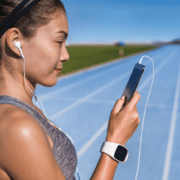 A woman standing on a blue running track, wearing a gray tank top, a white smartwatch, and white earphones, looks at her smartphone with a slight smile. The sky is clear and the background features an expansive green field. She's just finished pushing through the hard miles of her run. Austin Marathon Half Marathon & 5K