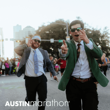 Two people dressed in suits and ties participate in a marathon. One, wearing a green jacket and sunglasses, points playfully at the camera, while the other, in a gray jacket and headband, smiles and waves. In the background are a crowd and city buildings. The text "AUSTINmarathon" appears at the bottom, dispelling running myths. Austin Marathon Half Marathon & 5K