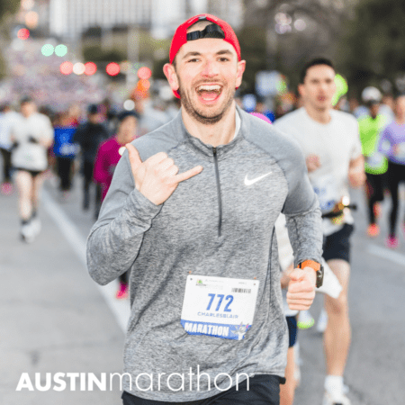 A smiling person in a gray long-sleeve shirt and red cap runs in the Austin Marathon amidst other runners. They hold their hand in a "shaka" sign and wear a race bib number 772 labeled "Charles Blair." The scene beautifully captures the spirit of marathon running, with an "Austin Marathon" logo on the image. Austin Marathon Half Marathon & 5K