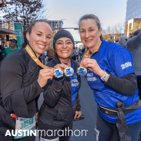 Three smiling marathon runners proudly display their medals after completing the Austin Marathon. Dressed in running attire, they stand together in a celebratory pose. A "PACE 3:25" sign and other participants are visible in the background, debunking common marathon myths. The text "Austin Marathon" is at the bottom. Austin Marathon Half Marathon & 5K