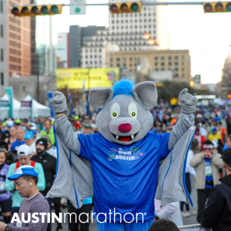 A large, costumed character dressed as a gray bat with a blue shirt that reads "Run Austin" raises its arms excitedly among a crowd of marathon runners in a city street. The text "AUSTIN marathon" is visible at the bottom of the image, capturing the vibrant spirit of marathon running. Austin Marathon Half Marathon & 5K