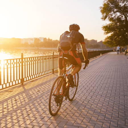 A cyclist rides along a paved path next to a body of water during sunset, likely integrating this ride into their marathon training. The person is wearing a helmet, backpack, and cycling gear. The path lined with a railing and trees hosts a few people in the background, all bathed in warm, golden light. Austin Marathon Half Marathon & 5K