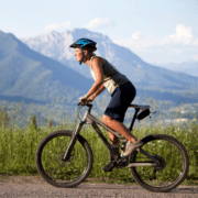 A person wearing a helmet rides a mountain bike on a gravel path, staying active and focused on their marathon goals. They are outdoors with tall grass by the roadside and a scenic view of mountains and a clear sky in the background. The rider is dressed in a sleeveless shirt and shorts. Austin Marathon Half Marathon & 5K