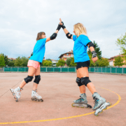 Two women wearing protective gear and rollerblades high-five each other outdoors on a paved area, showing their commitment to staying active. Both are dressed in blue t-shirts, with one in pink shorts and the other in black. Trees and buildings are visible in the background. Austin Marathon Half Marathon & 5K