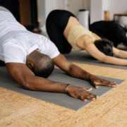 Three people are in a yoga class performing the child's pose on their mats. The room has a wooden floor, and the individuals are in comfortable clothing, stretching their arms forward with their foreheads touching the ground—prepping mind and body to stay active for marathon goals ahead. Austin Marathon Half Marathon & 5K