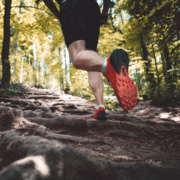 Close-up view of a person running uphill on a rocky, forested trail. The runner is wearing black shorts and red running shoes. Sunlight filters through the trees, casting dappled shadows on the path as they pursue their marathon goals. Austin Marathon Half Marathon & 5K