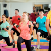 A group of people in a fitness class performing lunges with dumbbells, all aiming to achieve their marathon goals. They are on colorful exercise mats in a gym setting, dressed in athletic wear, and focusing on their workout in front of a large mirror. Austin Marathon Half Marathon & 5K