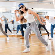 A group of individuals is dancing in a studio with wooden floors and mirrored walls, channeling their marathon goals. The center dancer, wearing a blue cap, sunglasses, a white tank top, gray pants, and white sneakers, strikes a dynamic pose. Other dancers are in the background, also in motion. Austin Marathon Half Marathon & 5K