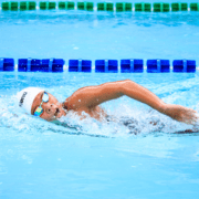 A swimmer, wearing a white swim cap and goggles, is performing the freestyle stroke in a pool. They are surrounded by blue and green lane dividers, creating a vibrant and focused scene of competitive swimming, highlighting the importance of staying active. Austin Marathon Half Marathon & 5K