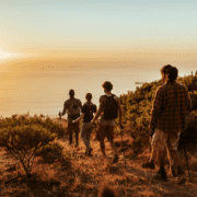 A group of five hikers with backpacks and trekking poles walk along a grassy trail overlooking the ocean at sunset, staying active and enjoying the outdoors. The sky is golden and the sea is calm, creating a serene and picturesque landscape. Austin Marathon Half Marathon & 5K