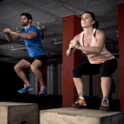 Two people are performing box jumps in a gym, staying active to achieve their marathon goals. The man on the left wears a blue shirt and black shorts, while the woman on the right sports a pink top and black leggings. Both are mid-air, jumping onto wooden boxes. Gym equipment and pillars fill the background. Austin Marathon Half Marathon & 5K