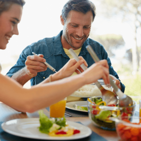 Two people sitting at a table, sharing a meal on bustling South Congress. The table is set with plates of fresh salad, a bowl of rice, and glasses of orange juice. One person serves salad while the other smiles, holding cutlery, ready to enjoy the food. Austin Marathon Half Marathon & 5K