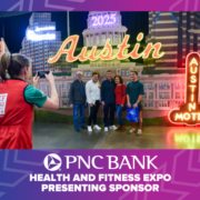 A group of people poses for a photo in front of an illuminated "Austin" sign at an indoor event featuring the Austin Marathon. The backdrop showcases the city's skyline, while a PNC Bank banner proudly reads, "Health and Fitness Expo Presenting Sponsor. Austin Marathon Half Marathon & 5K