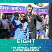 Two men wearing marathon medals stand smiling in front of a vendor backdrop for EIGHT Beer. They are posing happily, one holding a drink and giving a thumbs-up. The text reads, "The Official Beer of Austin Marathon," celebrating the exciting partnership. Austin Marathon Half Marathon & 5K