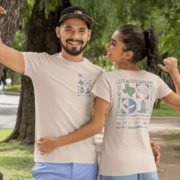 A smiling man and woman outdoors, each donning a light beige T-shirt with colorful graphics. The man's shirt proudly reads "Austin Marathon 2025," while the woman's back showcases vibrant Texas-themed designs. They pose playfully in a park setting, celebrating Race Day vibes. Austin Marathon Half Marathon & 5K