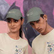Two people are wearing matching green caps and light beige Austin Marathon t-shirts. The person on the left sports a race bib numbered 2025, marking their excitement for Race Day as they stand against a colorful, abstract mural background. Austin Marathon Half Marathon & 5K