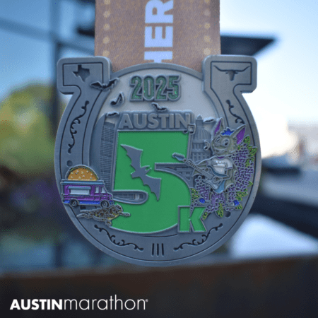 Close-up of a silver finisher medal for the Austin 5K 2025, featuring a bat, buildings, a moonlit sky, and lavender flowers. A food truck and a guitar add local flair to this race bling. The medal is suspended, with "Austin Marathon" printed in the corner. Austin Marathon Half Marathon & 5K