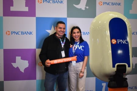 Two people smile for a photo in front of a PNC Bank-themed backdrop. One holds a PNC Bank foam stick, both wear event badges, and a photo booth stands nearby—showcasing the excitement of those who become a sponsor. Austin Marathon Half Marathon & 5K