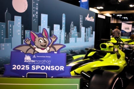 A cartoon bat mascot stands on a "2025 Sponsor" sign for the Austin Marathon, with a cityscape backdrop and a bright yellow-green race car—showcasing how you can become a sponsor and join the excitement. Austin Marathon Half Marathon & 5K