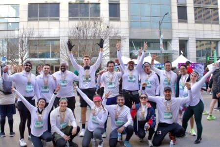 A group of marathon runners wearing white shirts and medals pose excitedly together outdoors, raising their hands and smiling after finishing a race—showcasing team management skills—with city buildings in the background. Austin Marathon Half Marathon & 5K