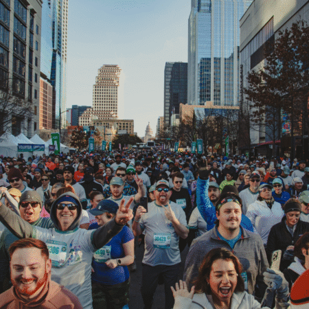 A large crowd of runners, many smiling and cheering, fills a city street during the Austin Marathon in May. Skyscrapers line both sides, and a domed building is visible in the distance under a clear sky as participants commit to the challenge. Austin Marathon Half Marathon & 5K
