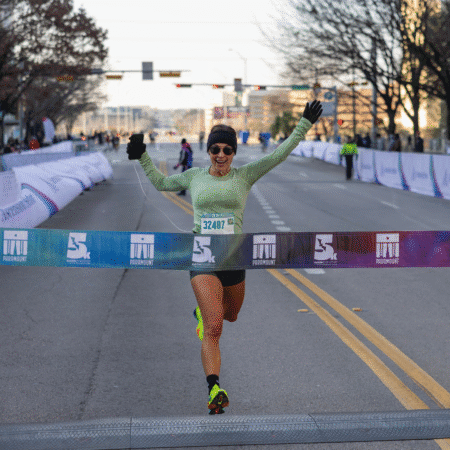 A runner wearing a green shirt and sunglasses crosses the finish line with arms raised in victory during the 2026 Austin Marathon on a city street. Austin Marathon Half Marathon & 5K