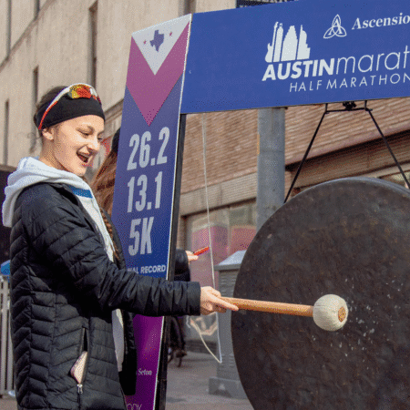 A woman in athletic clothing smiles while striking a large gong with a mallet at the Austin Marathon event, providing more reasons to get excited for 2026. A race sign showing distances is visible in the background. Austin Marathon Half Marathon & 5K