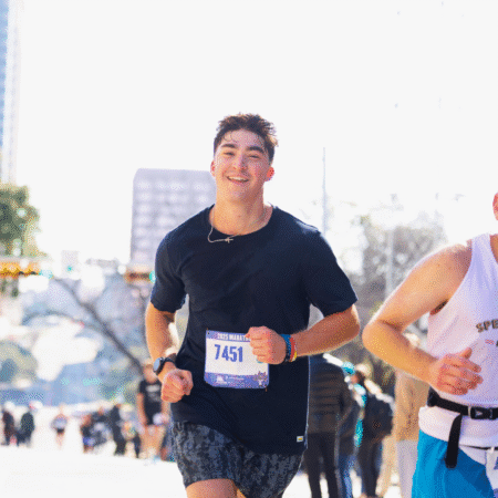 A young man wearing a race bib numbered 7451 runs in a city marathon, smiling and wearing a black t-shirt. Other runners from his training base and blurred city buildings are visible in the bright, sunny background. Austin Marathon Half Marathon & 5K