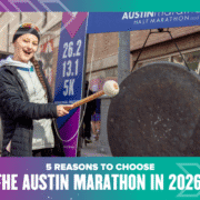 A smiling runner wearing a medal strikes a large gong at an outdoor event. Race distance signs for 26.2, 13.1, and 5K are visible. Text: "5 reasons to choose the Austin Marathon in 2026. Austin Marathon Half Marathon & 5K