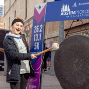 A smiling person in outdoor gear holds a mallet and stands next to a large gong at the Austin Marathon, ready to commit to their race goals in May, with a sign listing race distances behind them. Austin Marathon Half Marathon & 5K