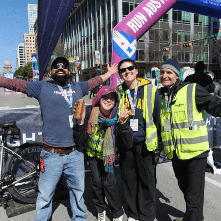 Four smiling adults, three in yellow safety vests and one in a blue shirt, pose together behind the scenes at an outdoor race event. One flashes a peace sign, and a “Run Austin” banner hints at the citywide celebration of the Austin Marathon. Austin Marathon Half Marathon & 5K