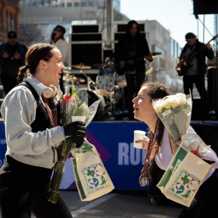 Two women with medals and flowers smile and celebrate after the Austin Marathon, holding drinks and gift bags, with a live band performing on a stage in the background during this citywide celebration. Austin Marathon Half Marathon & 5K