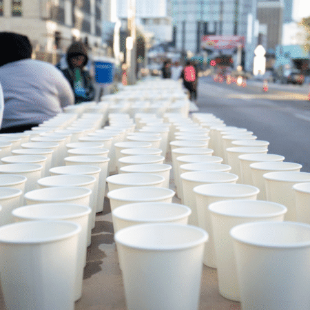 Rows of white paper cups filled with water are arranged on a table outdoors, ready for thirsty runners at the Austin Marathon—offering a behind-the-scenes glimpse of this citywide celebration, with blurred people and buildings in the background. Austin Marathon Half Marathon & 5K