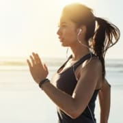 A woman with a ponytail jogs by the beach at sunrise, wearing a sleeveless top and earphones. Sunlight glows behind her, highlighting her focused expression as she tracks running calories burned on her fitness app. Austin Marathon Half Marathon & 5K