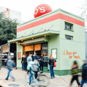 People walk and stand outside a green building with a red stripe and large red letters "O's" on the roof. Perfect for a day trip, the wall features the handwritten phrase "i love you so much," as others ride bikes past the entrance. Austin Marathon Half Marathon & 5K