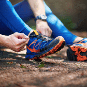 A person in blue athletic leggings sits on the ground outdoors in Austin, tying the orange and blue laces of a running shoe—a perfect scene for any beginner's guide to trail running, with leaves and dirt visible on the path. Austin Marathon Half Marathon & 5K