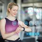 A woman in workout clothes checks her smartwatch at the gym. Digital graphs and circular stats show calories burned running, with percentages and data, plus multimedia control icons below. The background features blurred gym equipment. Austin Marathon Half Marathon & 5K