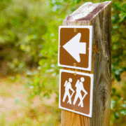 A wooden post displays two brown signs: one with a white left-pointing arrow above another showing two hikers with backpacks, indicating the direction of a hiking trail in Austin. Green foliage is blurred in the background—a great spot for trail running. Austin Marathon Half Marathon & 5K