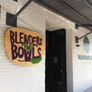 A storefront with a large wooden sign reading "Blenders and Bowls" in colorful letters is mounted on a white brick wall. The wall also displays the "Wanderlust Live" logo, showcasing one of the top healthy Austin restaurants. Austin Marathon Half Marathon & 5K
