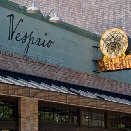 The exterior of a building in Austin with a sign reading “Vespaio” in cursive and a round yellow bee logo above it. The brick-and-glass facade and awning mark this spot as one of the city’s standout carb-loading restaurants. Austin Marathon Half Marathon & 5K