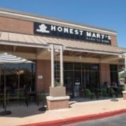 A restaurant storefront with a sign reading "Honest Mary's Farm to Bowl." Outdoor seating with green chairs and tables is visible under a large striped umbrella. Perfect for runners seeking healthy Austin restaurants, the brick building features large windows and a metal awning. Austin Marathon Half Marathon & 5K