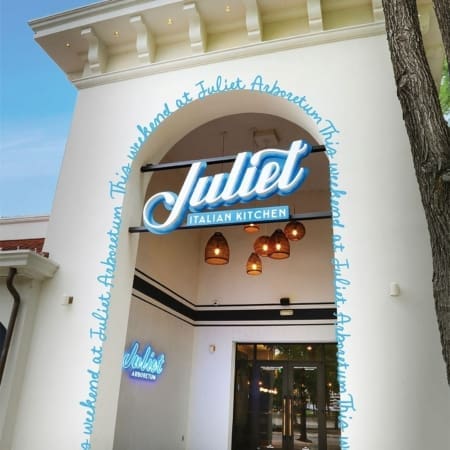 The entrance of Juliet Italian Kitchen in Austin features a large blue and white neon sign and hanging wicker lights, with "Juliet Arboretum" and a looping blue script text around the archway—a must-see among Austin restaurants. Austin Marathon Half Marathon & 5K