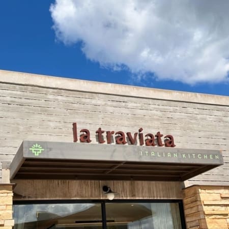 The exterior of a carb-loading restaurant in Austin features a sign reading "la traviata" and an awning labeled "Italian Kitchen" beneath a blue sky with clouds. Austin Marathon Half Marathon & 5K