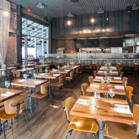 Modern Austin restaurant interior with wooden tables set with wine glasses and napkins, brown chairs, exposed brick walls, and an open kitchen in the background—a well-lit spot perfect for carb-loading restaurants enthusiasts. Austin Marathon Half Marathon & 5K